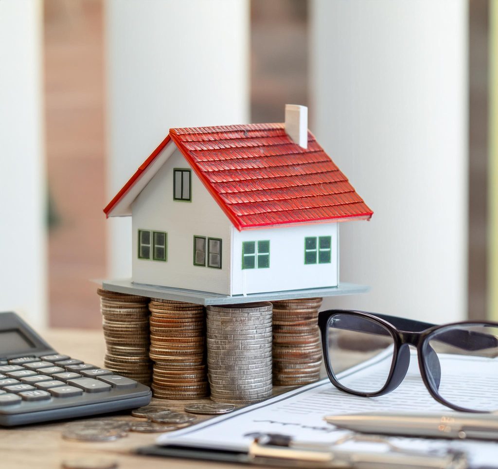 A porcelain miniature house model on coins, accompanied by a calculator, insurance forms, reading glasses, and dollar bills. The image represents themes of home ownership, financial planning, and insurance coverage. It conveys concepts related to real estate, insurance policies, and protecting assets