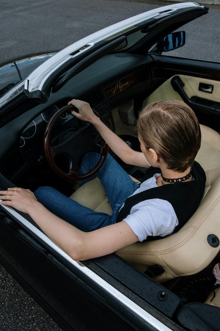 A person sitting in the driver's seat of a convertible car. The individual is holding the steering wheel with their right hand, while their left hand rests on the edge of the car's door. The person is dressed casually, wearing a white t-shirt, blue jeans, and a black vest. The interior of the car is a light beige color, and the convertible top is down, exposing the outside environment. The image is taken from an elevated angle, focusing on the person and the car's interior, conveying a sense of adulthood, freedom, style, and a relaxed driving experience.