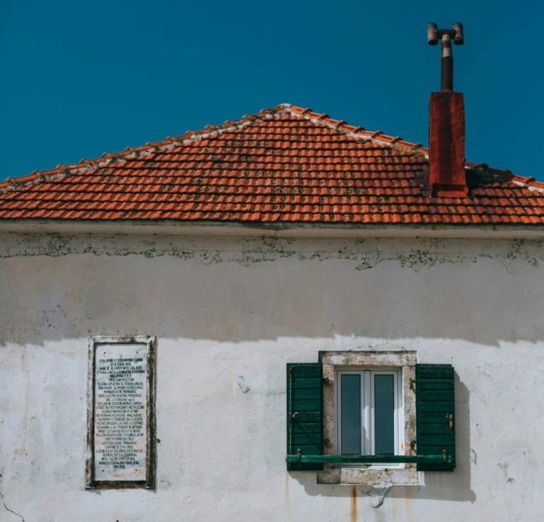 Front view of a weathered white building with a red tiled roof and green wooden shutters on the windows. The exterior shows signs of age, with visible cracks and stains on the walls. A wreath with a red ribbon hangs on the lower left side of the building, adding a touch of color. The rustic charm and character of the structure are highlighted against a clear blue sky, evoking a sense of history and simplicity.