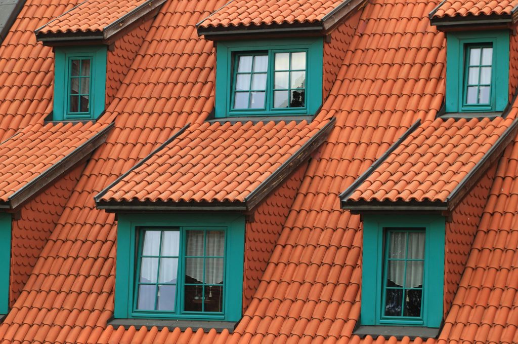 A close-up view of a roof with red-orange clay tiles and green-framed dormer windows. 