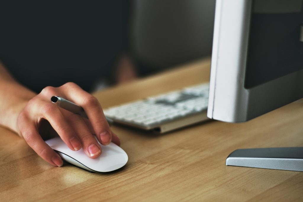 Close-up of a woman's hand using a wireless computer mouse, with a pen held between her fingers. The hand is positioned on a wooden desk in front of a desktop computer. In the background, a white keyboard is partially visible. The image conveys a sense of productivity, modern technology, and workplace activity. The focus is on the interaction between the user and the computer, highlighting the use of digital tools in a professional or home office setting.