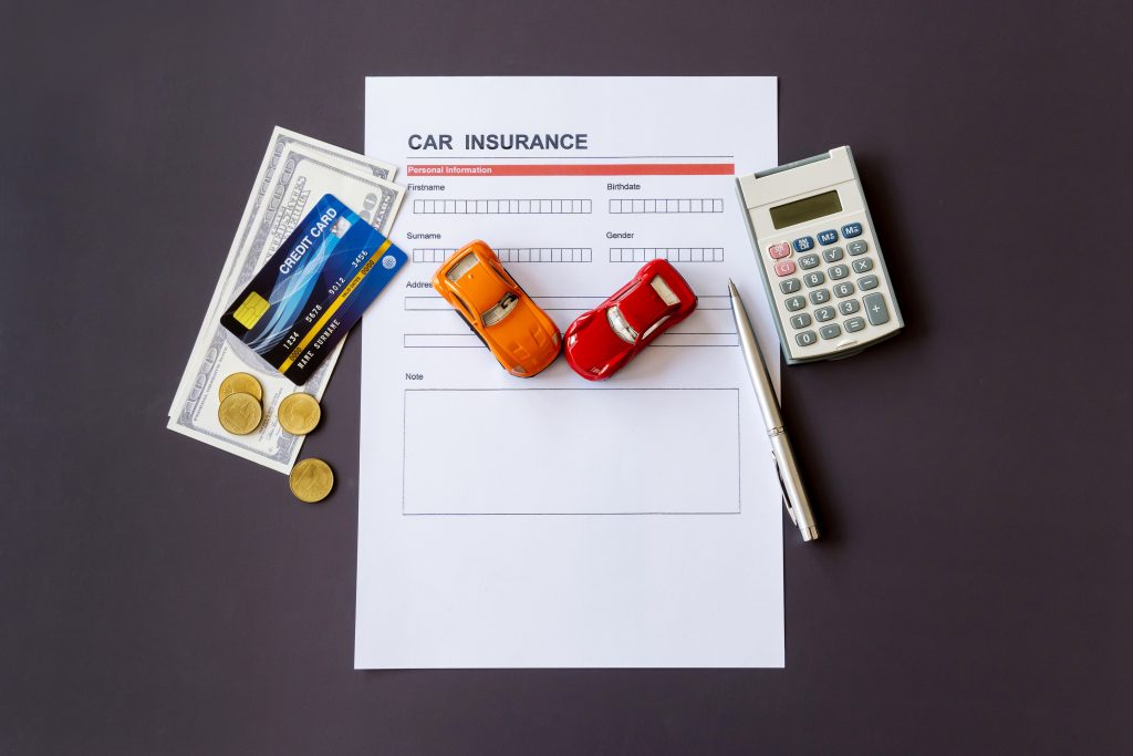 A flat lay of various items related to car insurance. At the center, a car insurance form, along with small toy cars. To the left of the form, a stack of cash, a credit card, and some coins are placed, indicating financial aspects. On the right side, a calculator and a pen are positioned, suggesting the calculation and completion of insurance details. The image conveys themes of financial planning, insurance, and automobile ownership, ideal for content related to car insurance, financial services, or automotive industry topics.