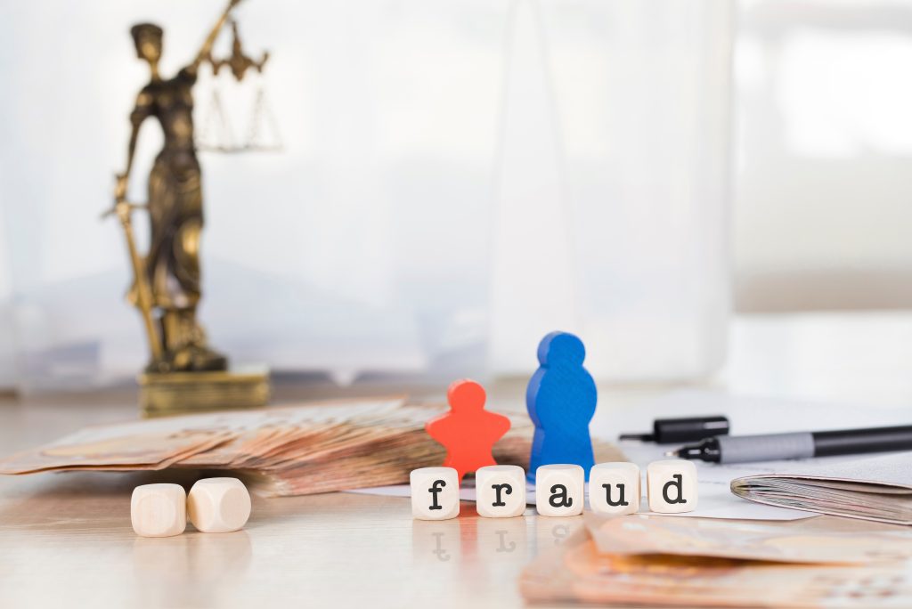 Close-up of the word 'fraud' spelled out with wooden letter blocks, placed on a desk with a blurry background of the Lady Justice statue, euro banknotes, and pens. The image symbolizes legal consequences, financial crime, and unethical practices, representing themes of fraud, justice, and law enforcement. Credit: Vecteezy