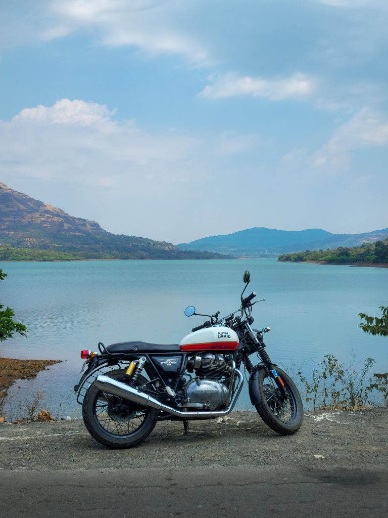 A Royal Enfield motorcycle parked by a scenic lakeside with mountains in the background.