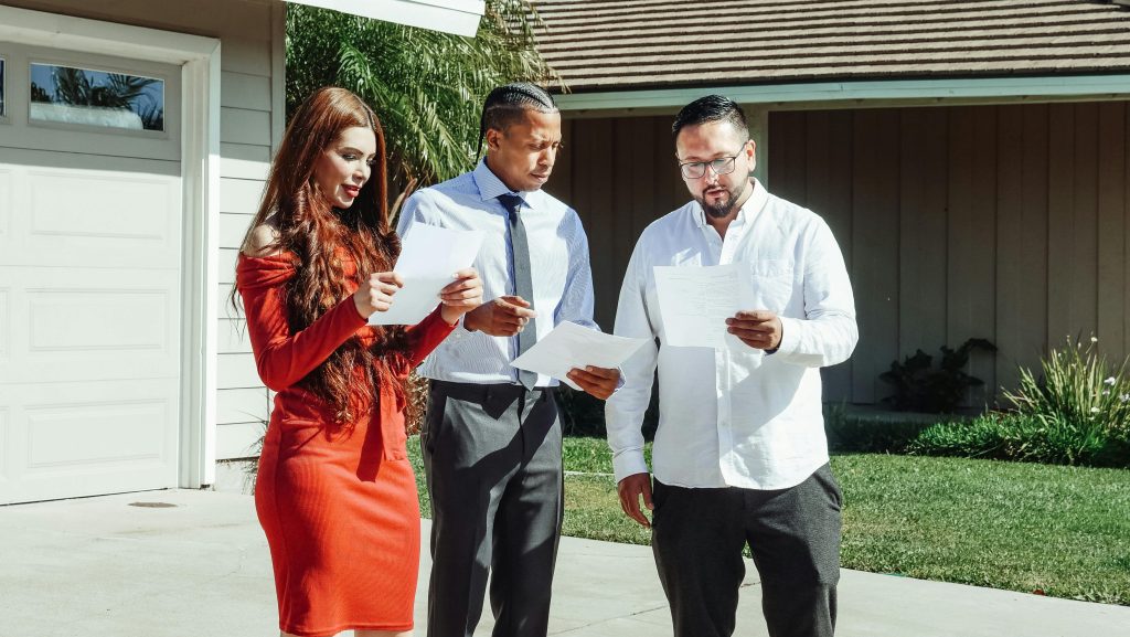 Three people are standing outdoors in front of a residential home, reviewing paperwork together. The group includes two men and one woman, all dressed professionally. The setting suggests a property or real estate consultation, possibly discussing insurance, mortgage documents, or a home inspection report. Concepts of "home insurance consultation," "homeowners", "real estate professionals," "property review"