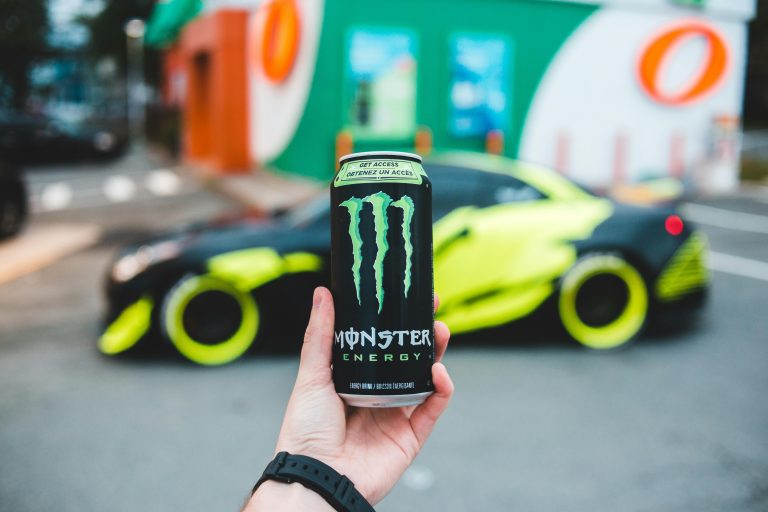 Close-up of a hand holding a can of Monster Energy drink with a blurred background of a neon yellow sports car and a vibrant urban setting.