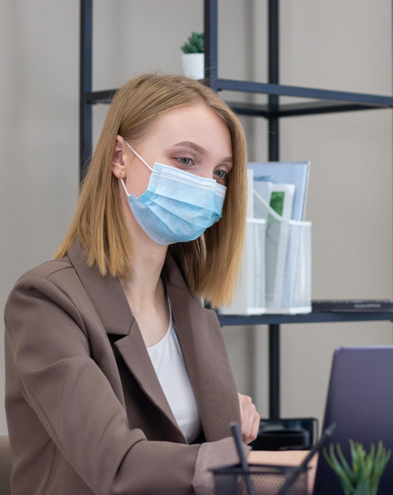 A professional woman wears a blue protective face mask while working on her laptop. The image reflects a safe and hygienic workplace or home office environment. Related to concepts of workplace safety, health precautions, paid sick leave, occupational health, or professional settings.