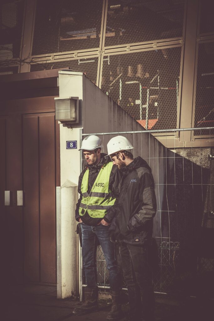 Two construction workers wearing hard hats, one in a reflective safety vest, stand near a building site, emphasizing the importance of pre-job safety evaluations and adherence to workplace safety standards for contractors