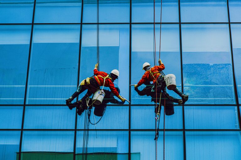 Two industrial workers in safety harnesses clean the glass facade of a high-rise building, highlighting the significance of proper training, equipment, and workplace safety measures for high-risk jobs in construction and maintenance