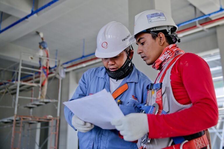 Two construction workers wearing safety gear review a document on-site, emphasizing safety planning and communication. A scaffolding structure and ongoing construction work are visible in the background, highlighting the importance of safety meetings and training in hazardous environments