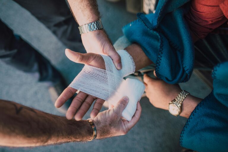 Close-up of a person receiving first aid for a hand injury, with another individual wrapping their hand in a gauze bandage. This image represents workplace injuries, workers' compensation claims, and the legal considerations of injuries occurring in parking lots or during work-related travel.